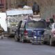 Man standing on top of a graffiti-covered trailer loaded with debris beside a blue minivan and a silver sedan on a city street; several people and a cart nearby (KOMO News).
