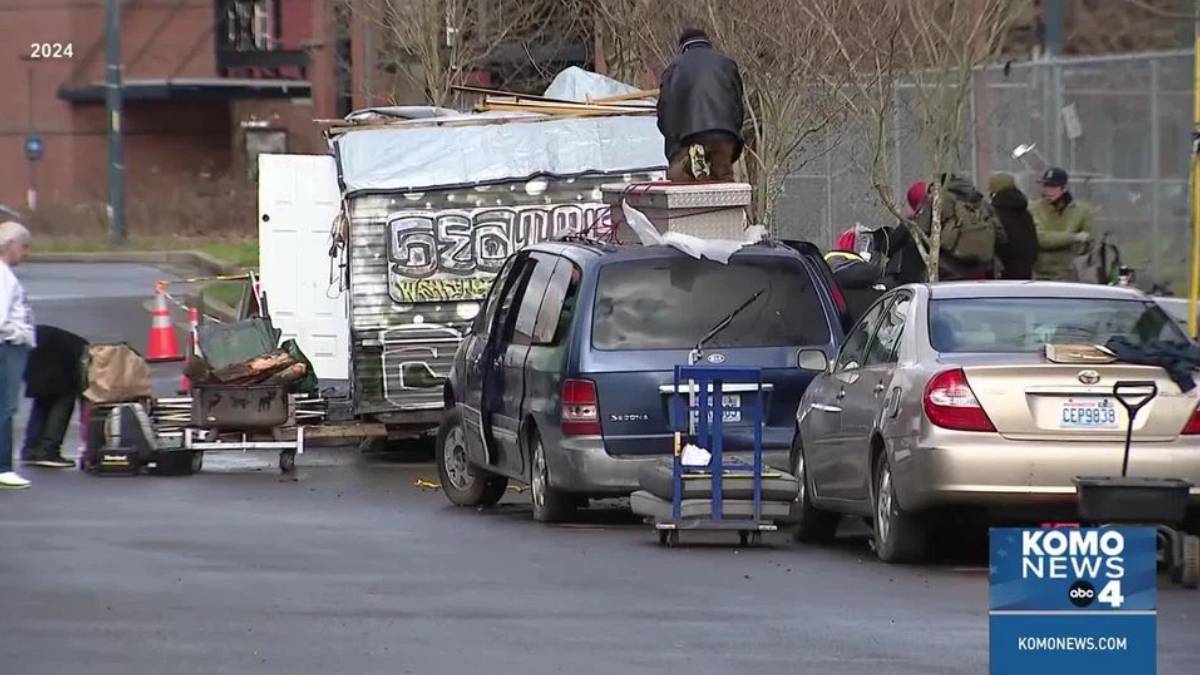 Man standing on top of a graffiti-covered trailer loaded with debris beside a blue minivan and a silver sedan on a city street; several people and a cart nearby (KOMO News).