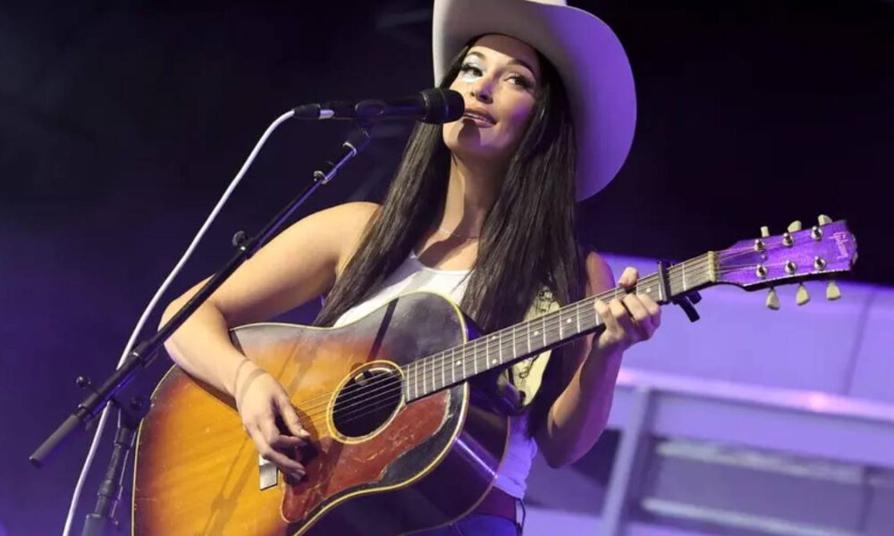 Female singer wearing a cowboy hat plays an acoustic guitar on stage and sings into a microphone.