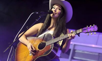 Female singer wearing a cowboy hat plays an acoustic guitar on stage and sings into a microphone.