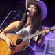 Female singer wearing a cowboy hat plays an acoustic guitar on stage and sings into a microphone.