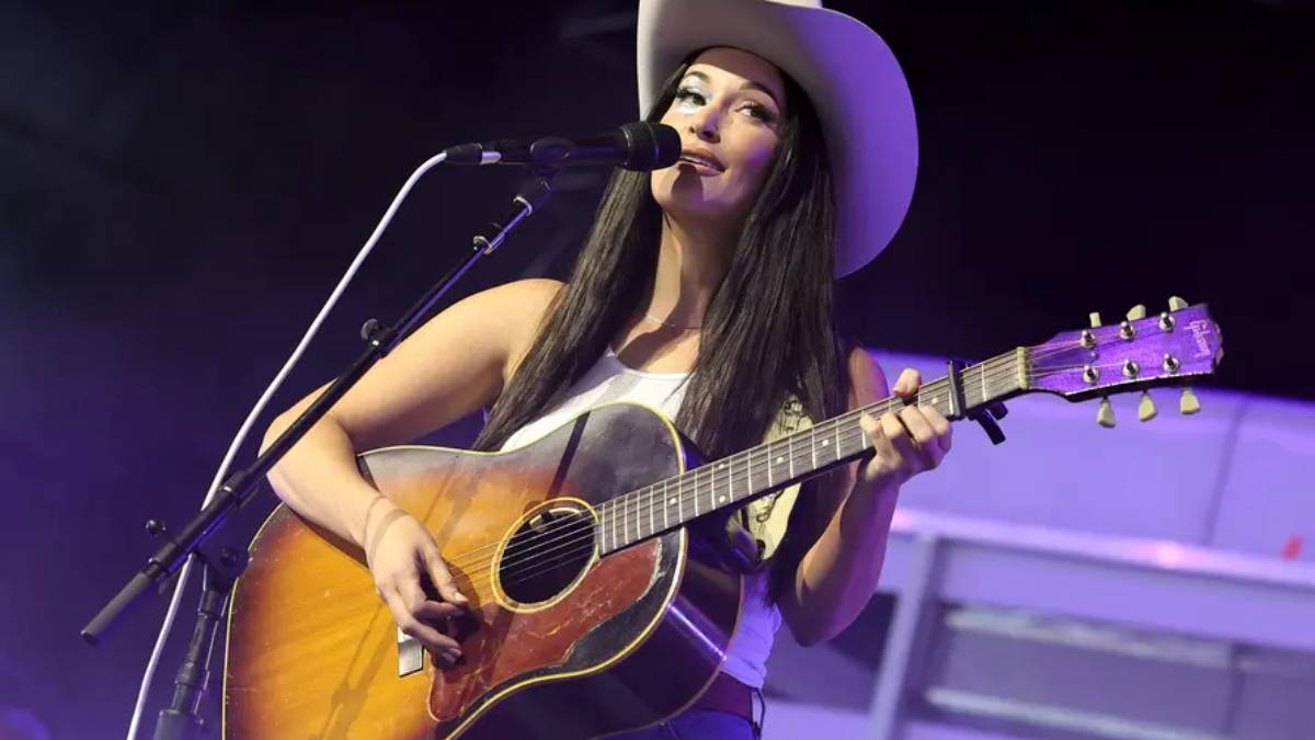 Female singer wearing a cowboy hat plays an acoustic guitar on stage and sings into a microphone.