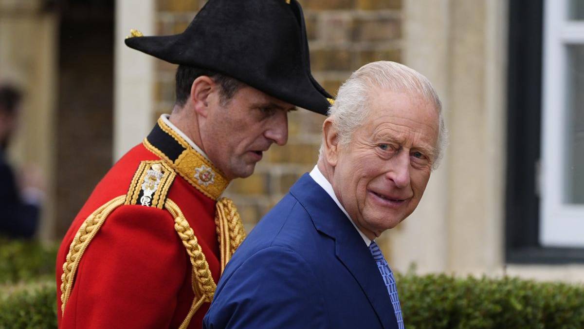 Older man in a blue suit and striped tie walking outdoors, with a ceremonial guard in a red uniform nearby.