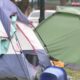 Colorful tents pitched on a grassy area near a street, forming an informal campsite: blue, green, and white tents visible side by side.