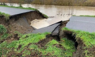 A road has collapsed into a large sinkhole, exposing soil and grass with muddy water pooling beyond the gap.