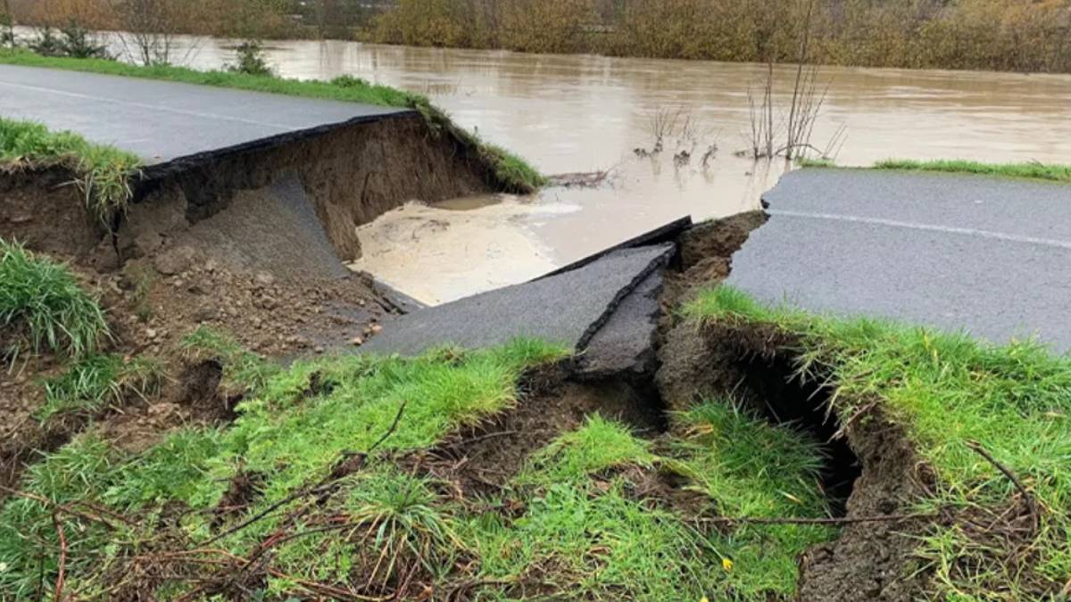 A road has collapsed into a large sinkhole, exposing soil and grass with muddy water pooling beyond the gap.