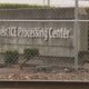 Chain-link fence in front of a sign that reads Northwest Processing Center behind a hedge-lined concrete wall.