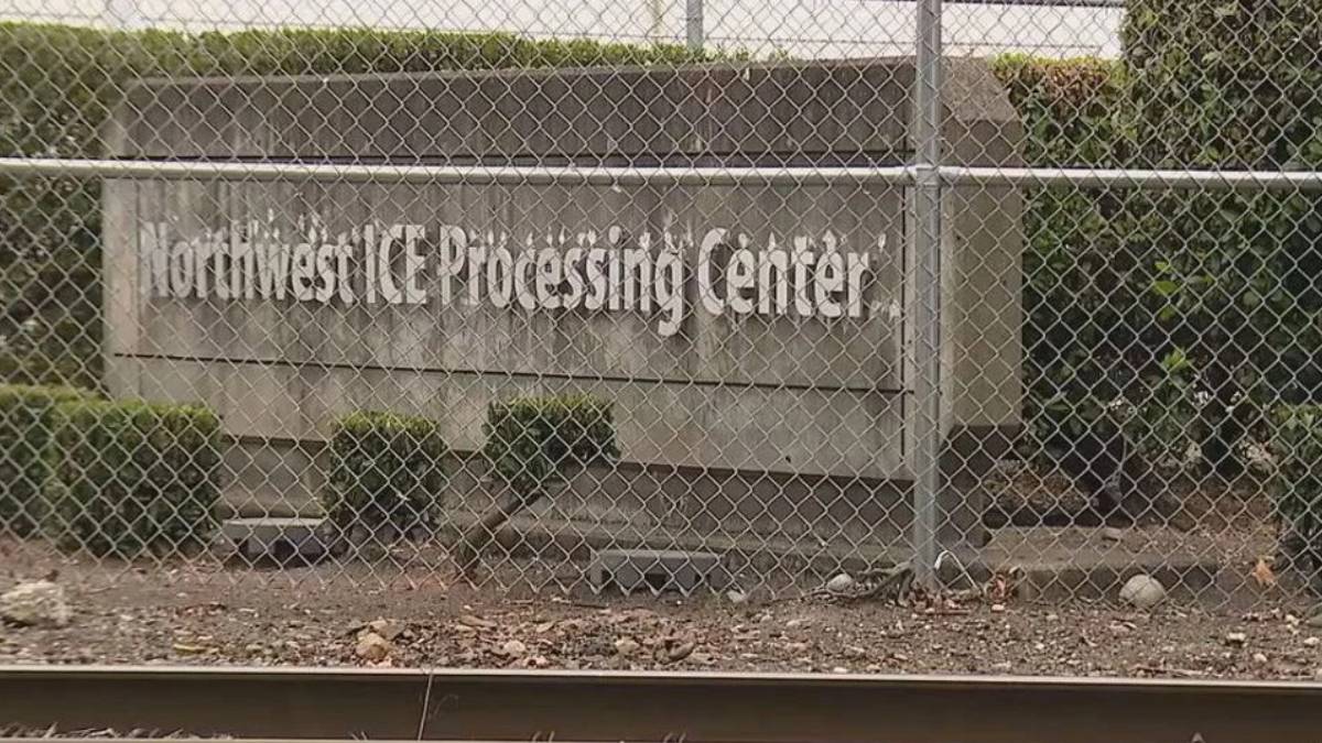 Chain-link fence in front of a sign that reads Northwest Processing Center behind a hedge-lined concrete wall.