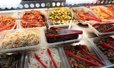 Buffet-style display of assorted prepared dishes in clear trays with red tongs, including noodles, greens, and meats.