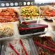Buffet-style display of assorted prepared dishes in clear trays with red tongs, including noodles, greens, and meats.