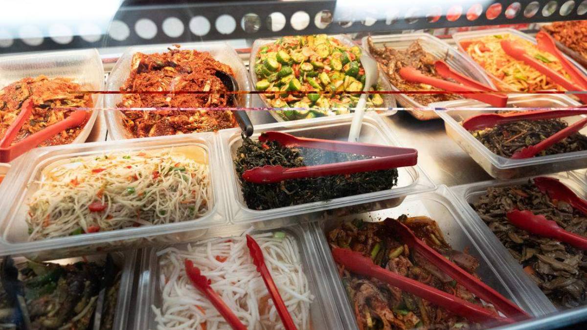 Buffet-style display of assorted prepared dishes in clear trays with red tongs, including noodles, greens, and meats.