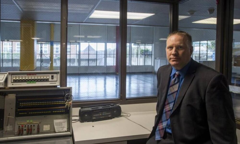 Man in a suit seated near vintage lab equipment with windows behind him in a tech room.