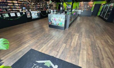 Interior of a cannabis shop with glass display cases, wall shelves stocked with products, and green accents along the walls and ceiling banners.