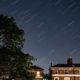 Night scene of a tree and brick houses under a star-trail sky with long-exposure star streaks above a quiet street.