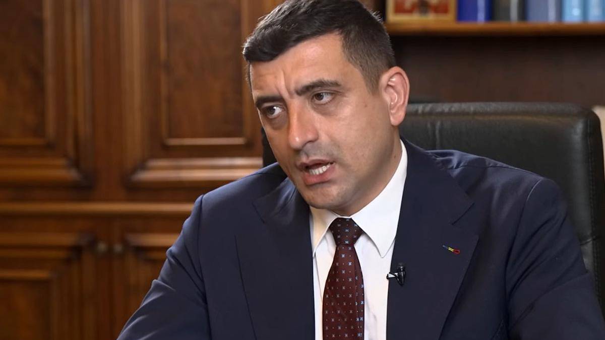 Man in a dark blue suit and maroon tie speaks in a formal office setting, with a microphone clipped to his lapel.