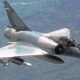 Grey military fighter jet in flight over a green landscape, single-seat cockpit visible and landing gear retracted, mid-depth perspective from above