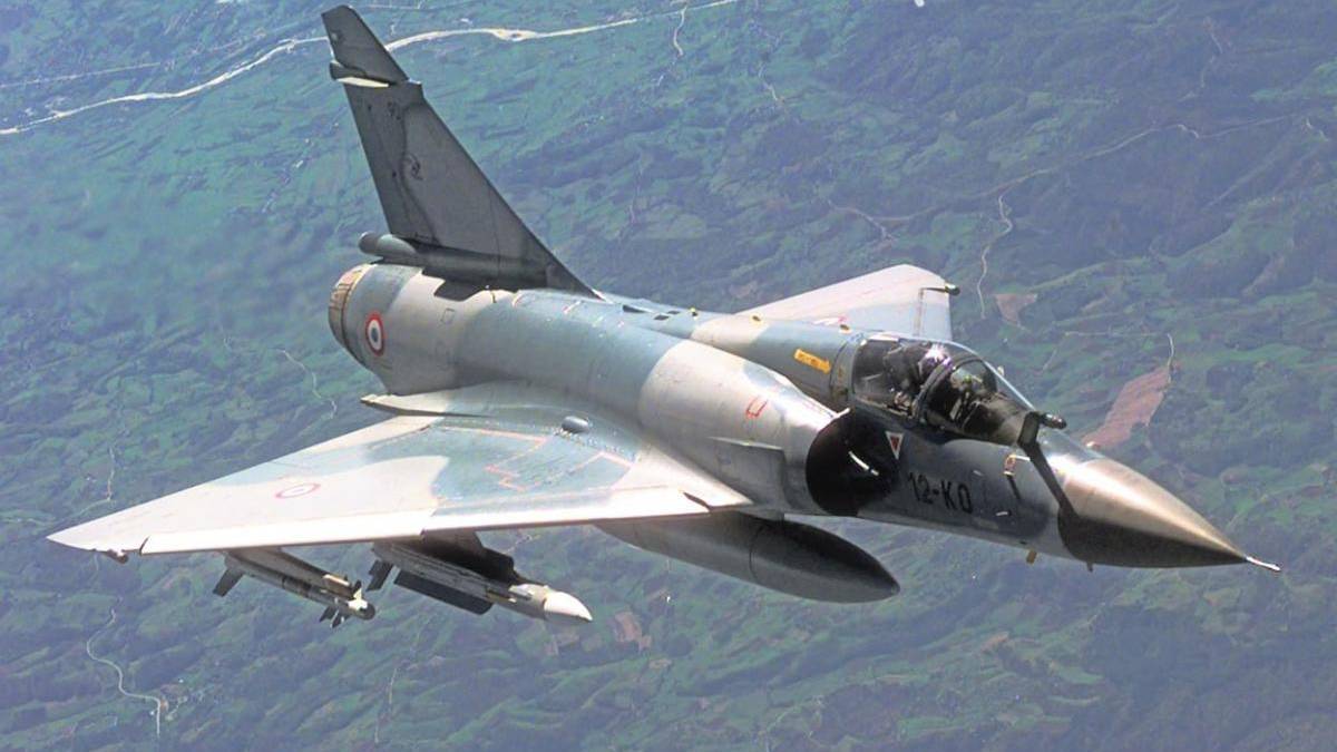 Grey military fighter jet in flight over a green landscape, single-seat cockpit visible and landing gear retracted, mid-depth perspective from above