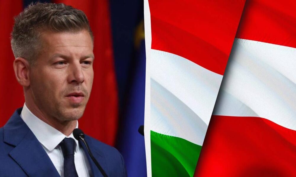 Man in a navy suit speaking at a microphone, with a red backdrop, and a Hungarian flag motif on the right side of the image.
