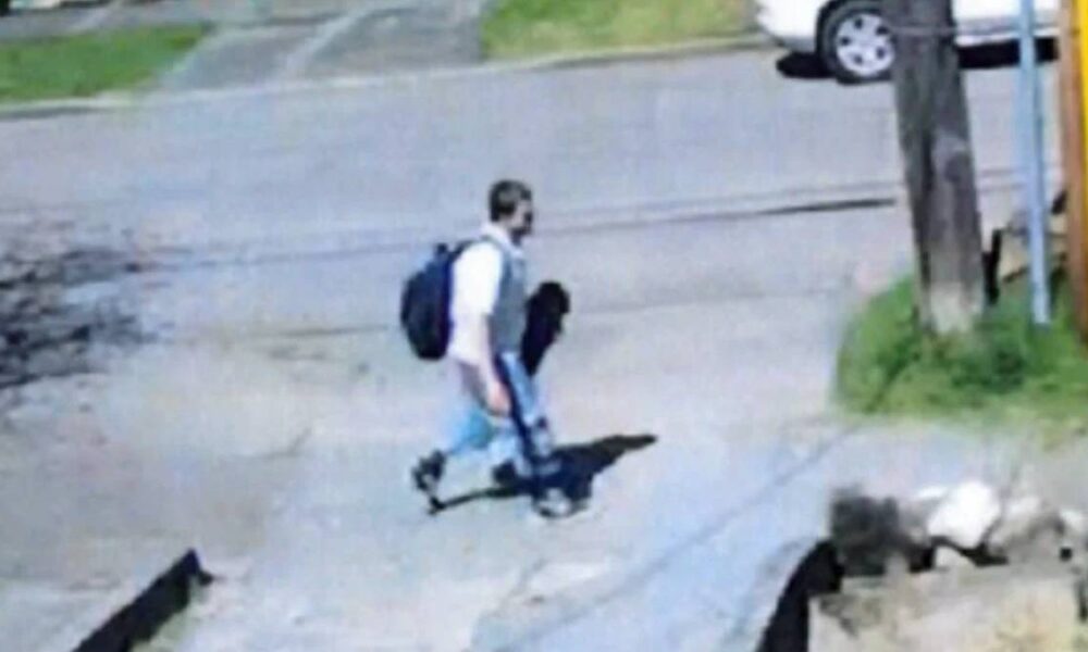 Young man with a backpack walking across a street curb in a residential area, sidewalk and parked cars visible in the background