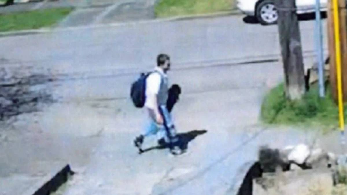Young man with a backpack walking across a street curb in a residential area, sidewalk and parked cars visible in the background
