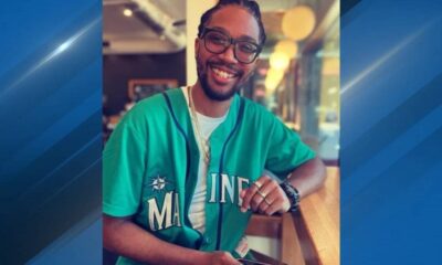 Smiling man with glasses and braided hair, wearing a teal Maine baseball jersey, seated at a cafe counter.