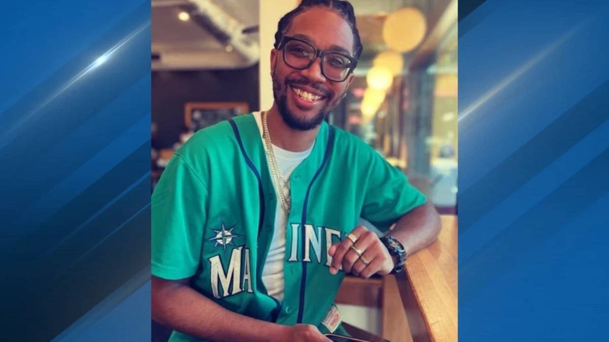 Smiling man with glasses and braided hair, wearing a teal Maine baseball jersey, seated at a cafe counter.