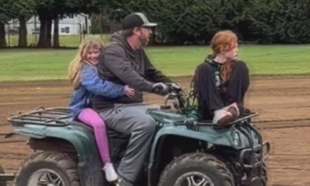 Man drives a green ATV on a dirt track with a blonde girl in a blue jacket sitting behind him and a red-haired girl sitting next to him on the front rack, outdoors near trees.