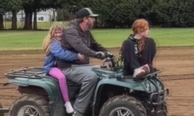 Man drives a green ATV on a dirt track with a blonde girl in a blue jacket sitting behind him and a red-haired girl sitting next to him on the front rack, outdoors near trees.