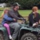 Man drives a green ATV on a dirt track with a blonde girl in a blue jacket sitting behind him and a red-haired girl sitting next to him on the front rack, outdoors near trees.