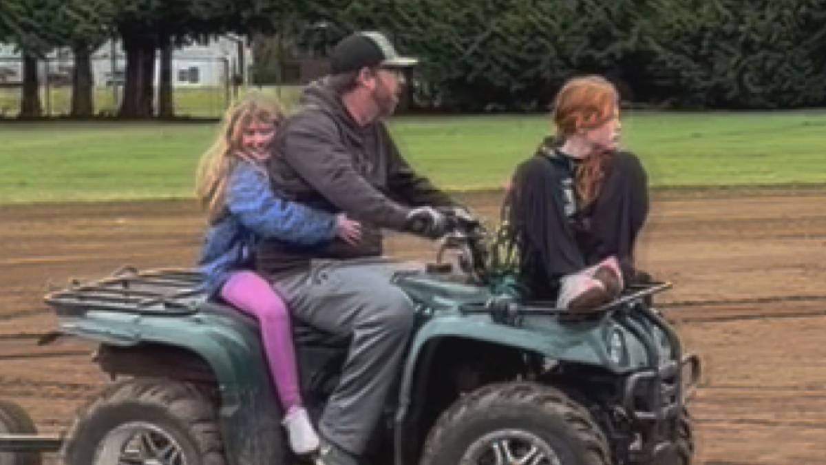 Man drives a green ATV on a dirt track with a blonde girl in a blue jacket sitting behind him and a red-haired girl sitting next to him on the front rack, outdoors near trees.