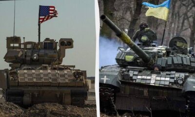 Two battle tanks side by side in a field, left with an American flag, right with a Ukrainian flag on their turrets.