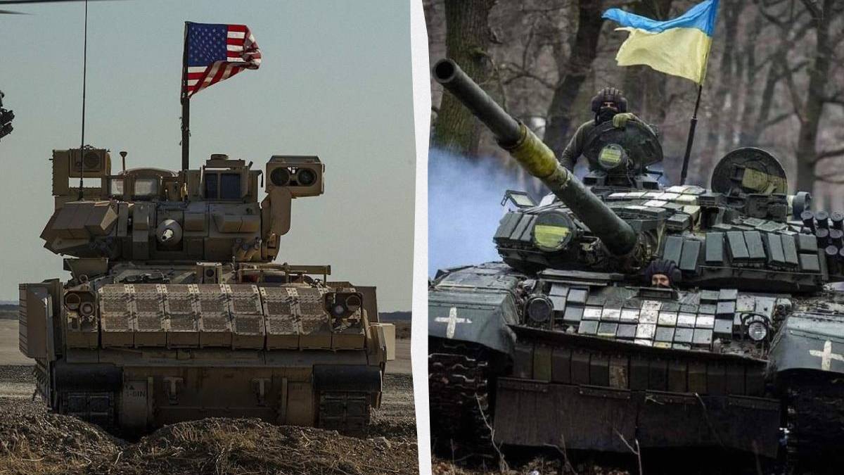 Two battle tanks side by side in a field, left with an American flag, right with a Ukrainian flag on their turrets.