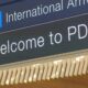 Airport sign reading 'International Arrivals' with airplane icon and 'Welcome to PDX'.