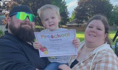 Family portrait: bearded man in black shirt and green sunglasses, woman in plaid shirt, and a toddler holding a 'Preschool Diploma' outside in a backyard park.