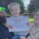 Family portrait: bearded man in black shirt and green sunglasses, woman in plaid shirt, and a toddler holding a 'Preschool Diploma' outside in a backyard park.