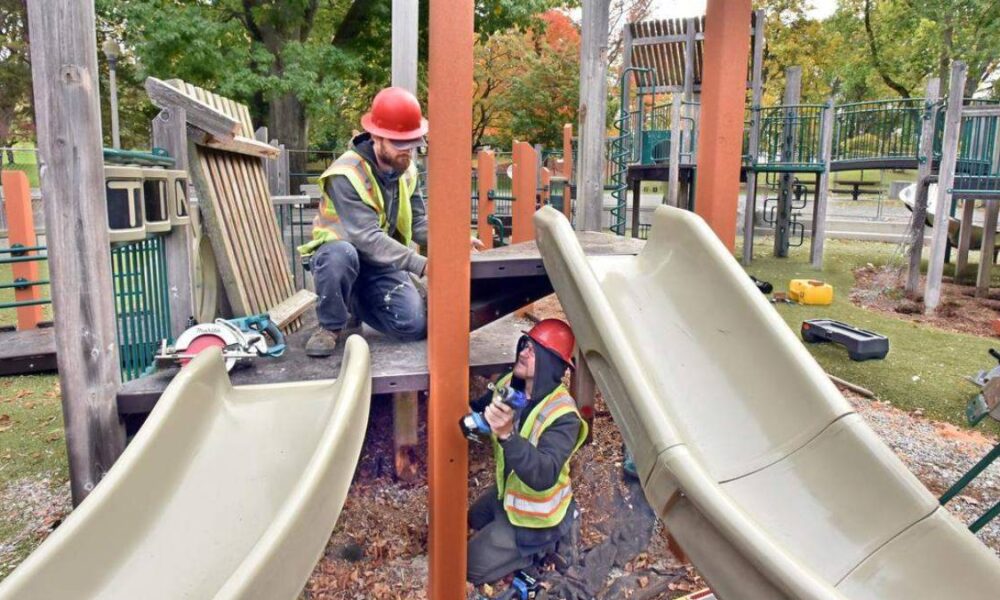Two construction workers in hard hats and reflective vests repairing a playground slide on a wooden platform.