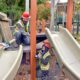 Two construction workers in hard hats and reflective vests repairing a playground slide on a wooden platform.