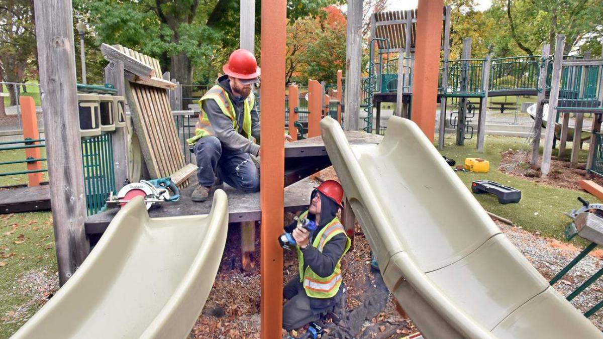 Two construction workers in hard hats and reflective vests repairing a playground slide on a wooden platform.