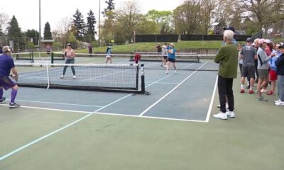 Doubles tennis match on an outdoor court with spectators lined up along the fence.
