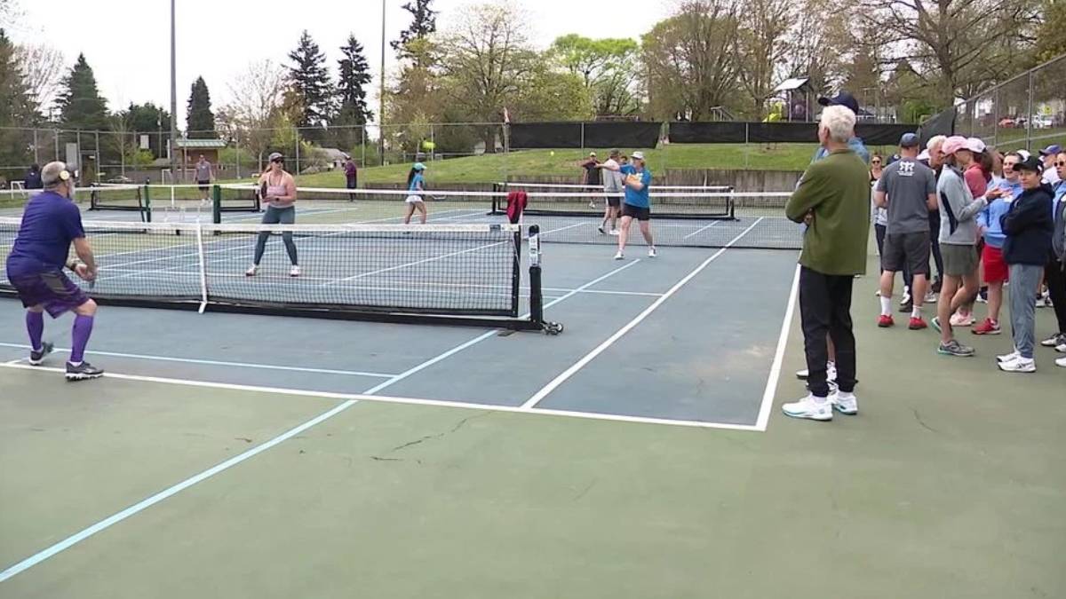 Doubles tennis match on an outdoor court with spectators lined up along the fence.