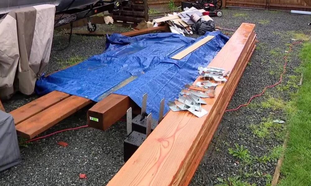 Outdoor work area with long wooden beams and a blue tarp spread over gravel, surrounded by scattered hardware and tools.