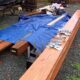 Outdoor work area with long wooden beams and a blue tarp spread over gravel, surrounded by scattered hardware and tools.