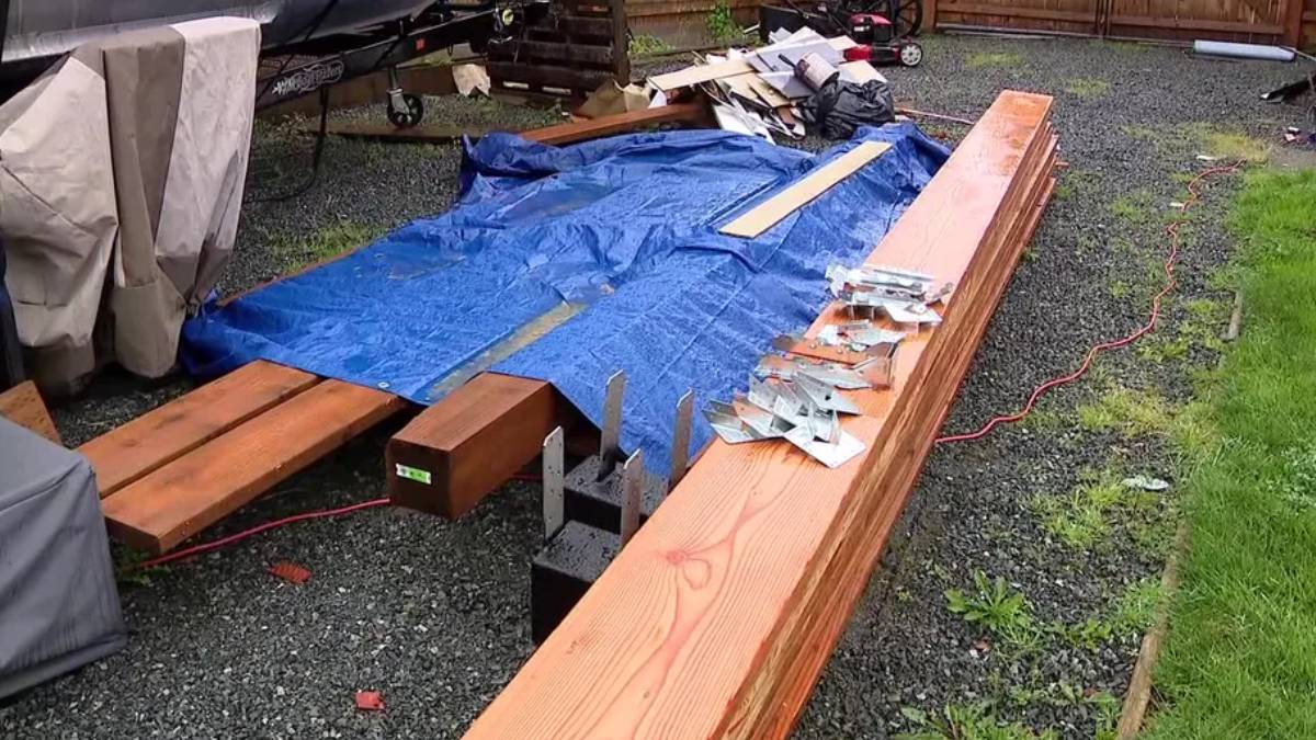 Outdoor work area with long wooden beams and a blue tarp spread over gravel, surrounded by scattered hardware and tools.