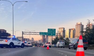 Highway scene with police SUV and orange traffic cones blocking lanes, city skyline in the distance.