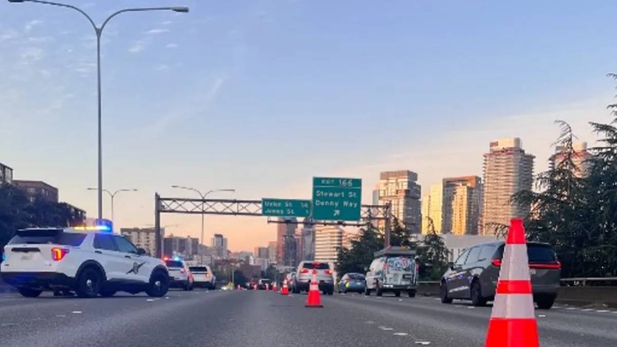 Highway scene with police SUV and orange traffic cones blocking lanes, city skyline in the distance.