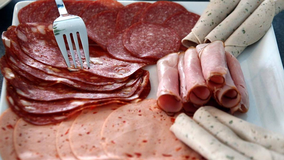 Assorted deli meats on a white platter: pepperoni slices, salami, rolled ham, and pale herb-infused sausages with a fork resting on top.