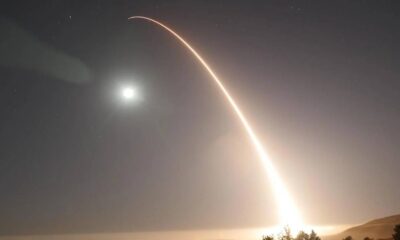 Long-exposure night photo of a bright rocket ascent leaving a curved orange trail across the sky, with the moon and trees on the horizon.