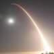 Long-exposure night photo of a bright rocket ascent leaving a curved orange trail across the sky, with the moon and trees on the horizon.