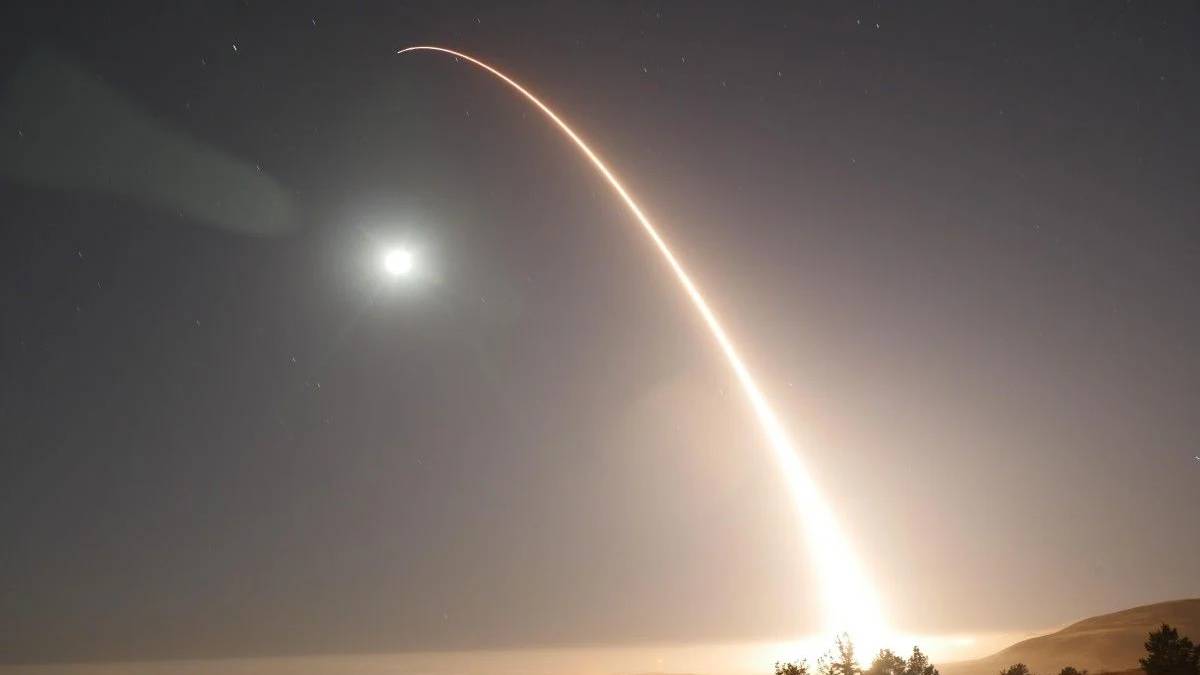 Long-exposure night photo of a bright rocket ascent leaving a curved orange trail across the sky, with the moon and trees on the horizon.