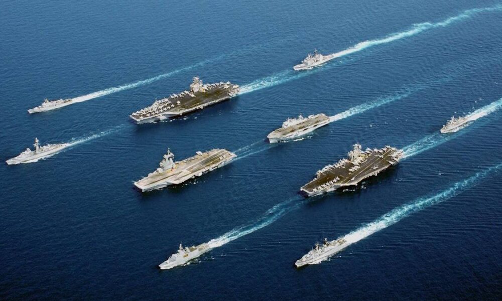 Formation of aircraft carriers and escort ships sailing in open blue ocean, leaving white wakes behind them.